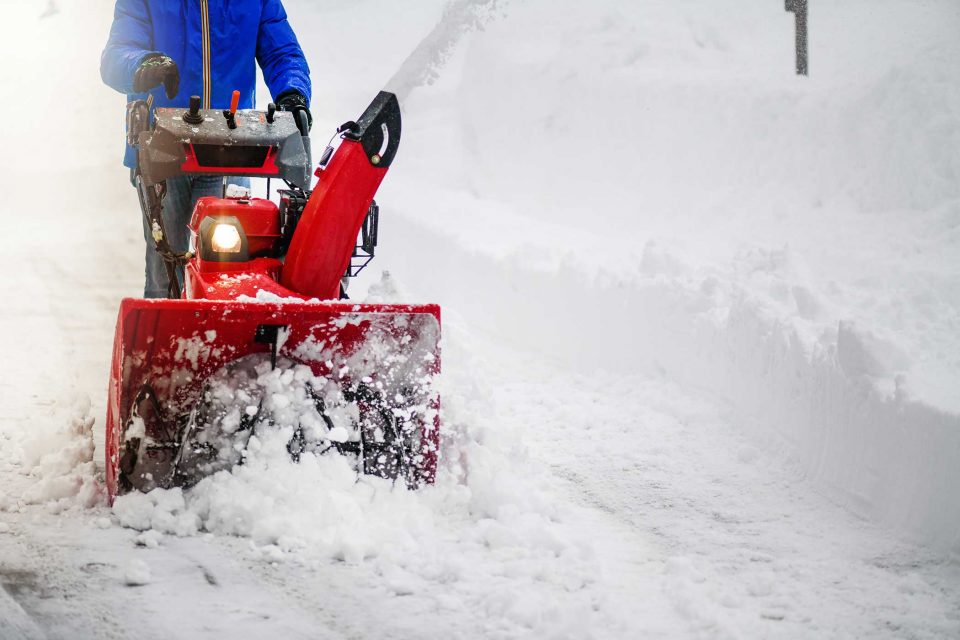 Person in blue jacket using a red snow blower for snow removal on a street.